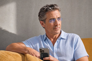 Man wearing ThinOptics Moment blue light blocking armless readers holding an iPhone with a ThinOptics Pod case on a yellow sofa