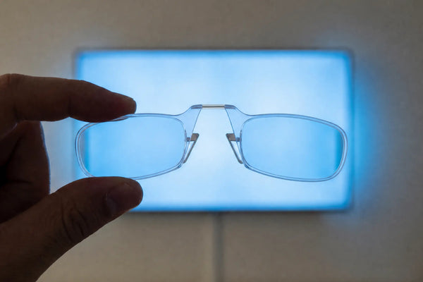 Hand holding ThinOptics Moment blue light blocking armless readers up against a glowing blue screen