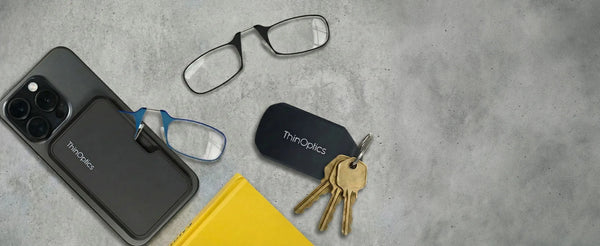 ThinOptics phone case with blue armless readers, black armless reading glasses, and a keychain case on keys arranged on a concrete surface with a yellow notebook