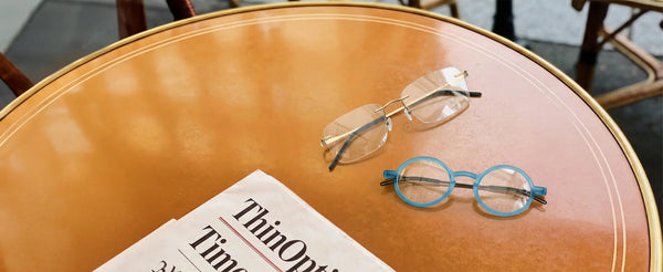 ThinOptics rimless gold and blue round prescription glasses resting on a warm-toned cafe table beside a ThinOptics newspaper