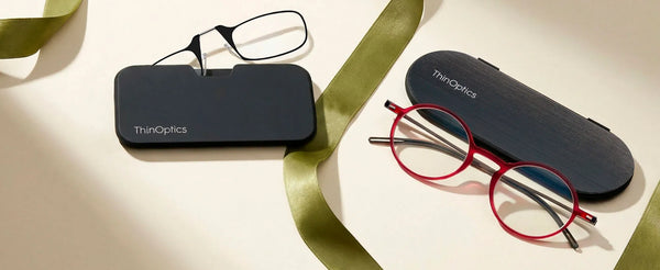 Two ThinOptics reading glasses with slim cases on a cream surface — black rectangular frames with a phone case, and red round frames with an oval case, surrounded by a gold ribbon