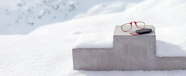 Red ThinOptics reading glasses and compact keychain case displayed on concrete block in snowy outdoor setting