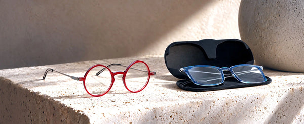 ThinOptics red round reading glasses beside blue rectangle readers resting in an open black Brooklyn case on a stone surface in warm sunlight