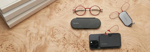 ThinOptics red round reading glasses, red armless readers on a phone case and keychain case, and a slim Milano oval case arranged on a warm burl wood surface