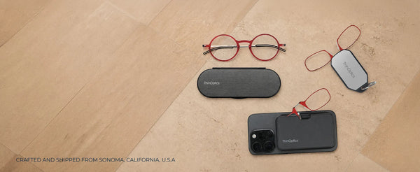 ThinOptics red round reading glasses, armless readers on a phone case and keychain case, and a slim Milano oval case arranged on a warm stone surface