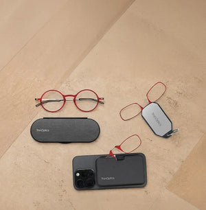 ThinOptics red round reading glasses, armless readers on a phone case and keychain case, and a slim Milano oval case arranged on a warm stone surface