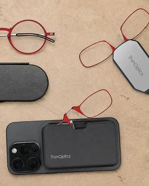 ThinOptics red round reading glasses, red armless readers on a phone case and keychain case, and a slim Milano oval case on a sandy stone surface