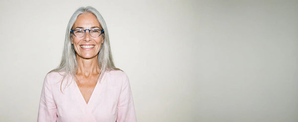 Smiling woman with long gray hair wearing ThinOptics blue cat-eye prescription glasses against a neutral background