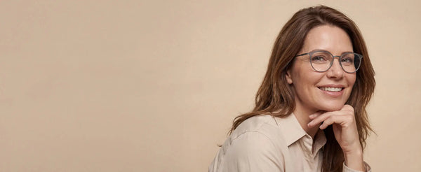 Smiling woman wearing ThinOptics round prescription glasses with her chin resting on her hand against a warm beige background
