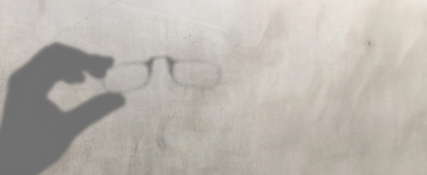 Shadow of a hand holding a pair of reading glasses cast across a worn concrete surface