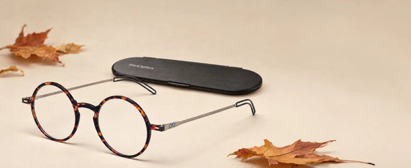 ThinOptics tortoise round reading glasses beside a black Brooklyn case surrounded by autumn maple leaves on a warm beige surface