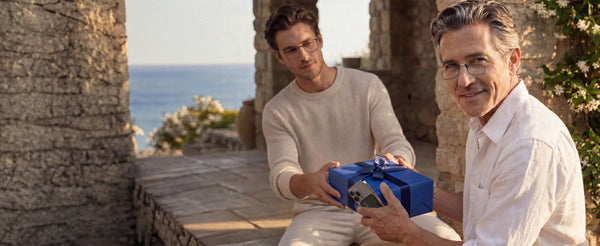 Two men wearing ThinOptics reading glasses exchanging a wrapped gift box on a stone terrace overlooking the sea