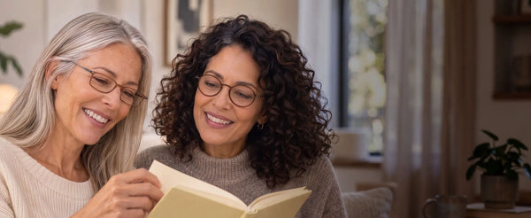 Two smiling women wearing ThinOptics reading glasses sharing a book together in a warm, cozy home setting