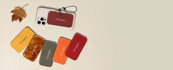 ThinOptics Universal Pod Cases in fall colors — yellow, burlwood, gray, orange, and red — with one attached to an iPhone and an autumn maple leaf