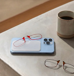 Red ThinOptics armless readers resting on a white Universal Pod attached to a blue iPhone, with tortoiseshell armless readers on a concrete surface nearby