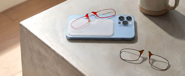 Red ThinOptics armless readers resting on a white Universal Pod attached to a blue iPhone, with tortoiseshell armless readers on a concrete surface nearby