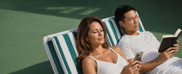 Woman and man both wearing ThinOptics red reading glasses relaxing on striped pool loungers, she on her phone and he reading a book