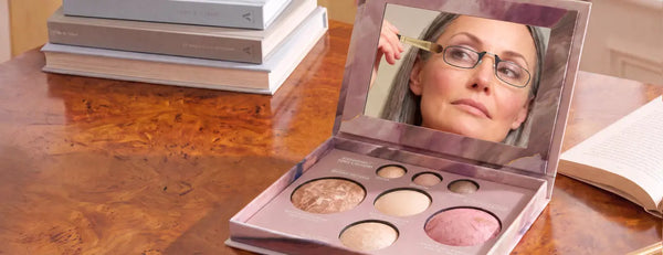 Woman wearing ThinOptics black reading glasses applying eye makeup, reflected in an open cosmetics compact on a wood table