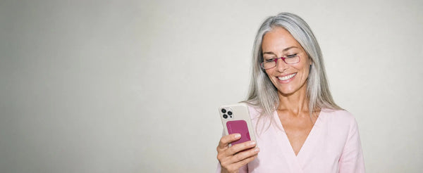 Smiling woman wearing ThinOptics pink armless reading glasses looking at her phone with a pink ThinOptics Pod case attached