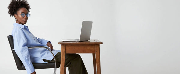 Woman wearing ThinOptics round blue light blocking glasses sitting at a wooden desk with a laptop on a white background