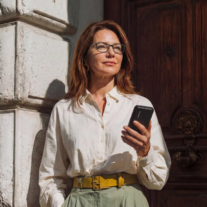 Woman in a white linen shirt wearing black rectangular reading glasses holding a phone with a ThinOptics case, leaning against a sunlit stone wall by a wooden door