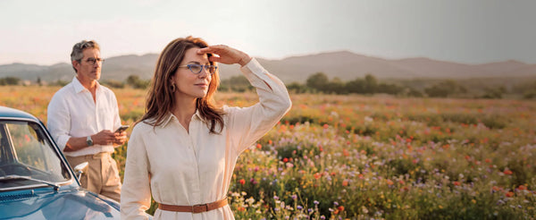 Woman in cat-eye reading glasses shielding her eyes while gazing across a colorful wildflower field, with a man wearing glasses standing by a vintage car behind her