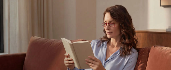 Woman in a blue blouse wearing red reading glasses relaxing on a terracotta couch reading a book in a warm sunlit living room